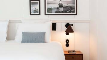 Una cama bien hecha con sábanas blancas y almohadas estampadas, una mesita de noche con lámpara, y dos cuadros en la pared.
