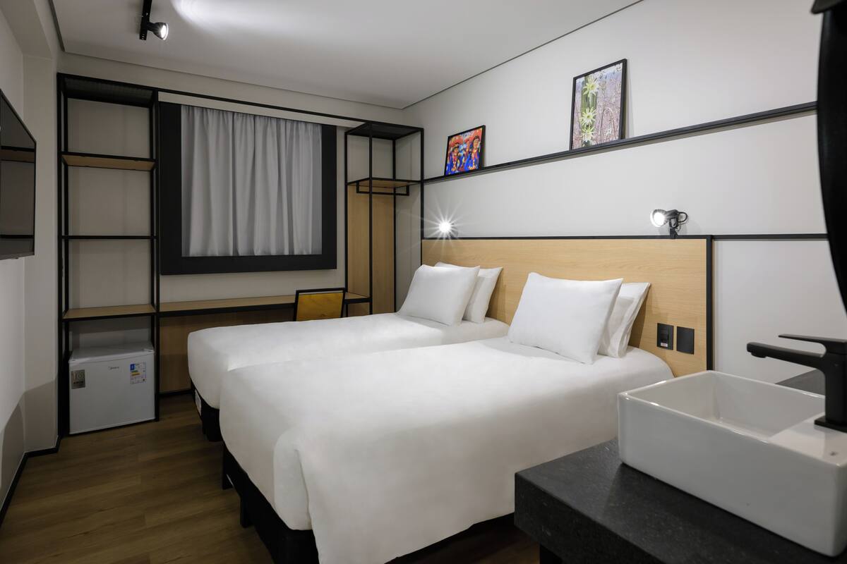 Ein Hotelzimmer mit zwei Betten, einem Schreibtisch, einer kleinen Kühlschrank, einem Nachttisch, einem Waschbecken und Bildern an der Wand.
