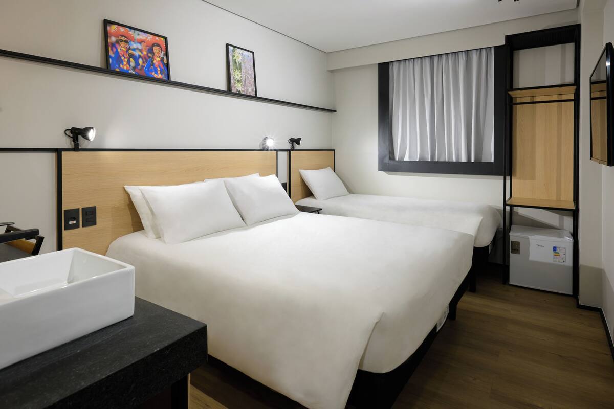 Ein Hotelzimmer mit zwei Betten, einem Schreibtisch, einem Stuhl, einem kleinen Tisch, einem Regal mit Kunstwerken, einem Fenster mit Vorhängen und einem Einbauschrank.
