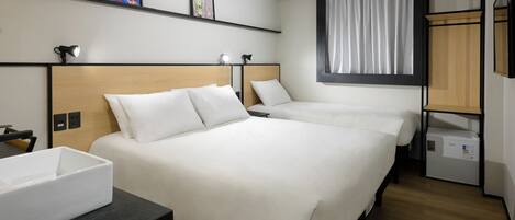 A hotel room with two beds, a desk, a chair, a small table, a shelf with artwork, a window with curtains, and a built-in wardrobe.