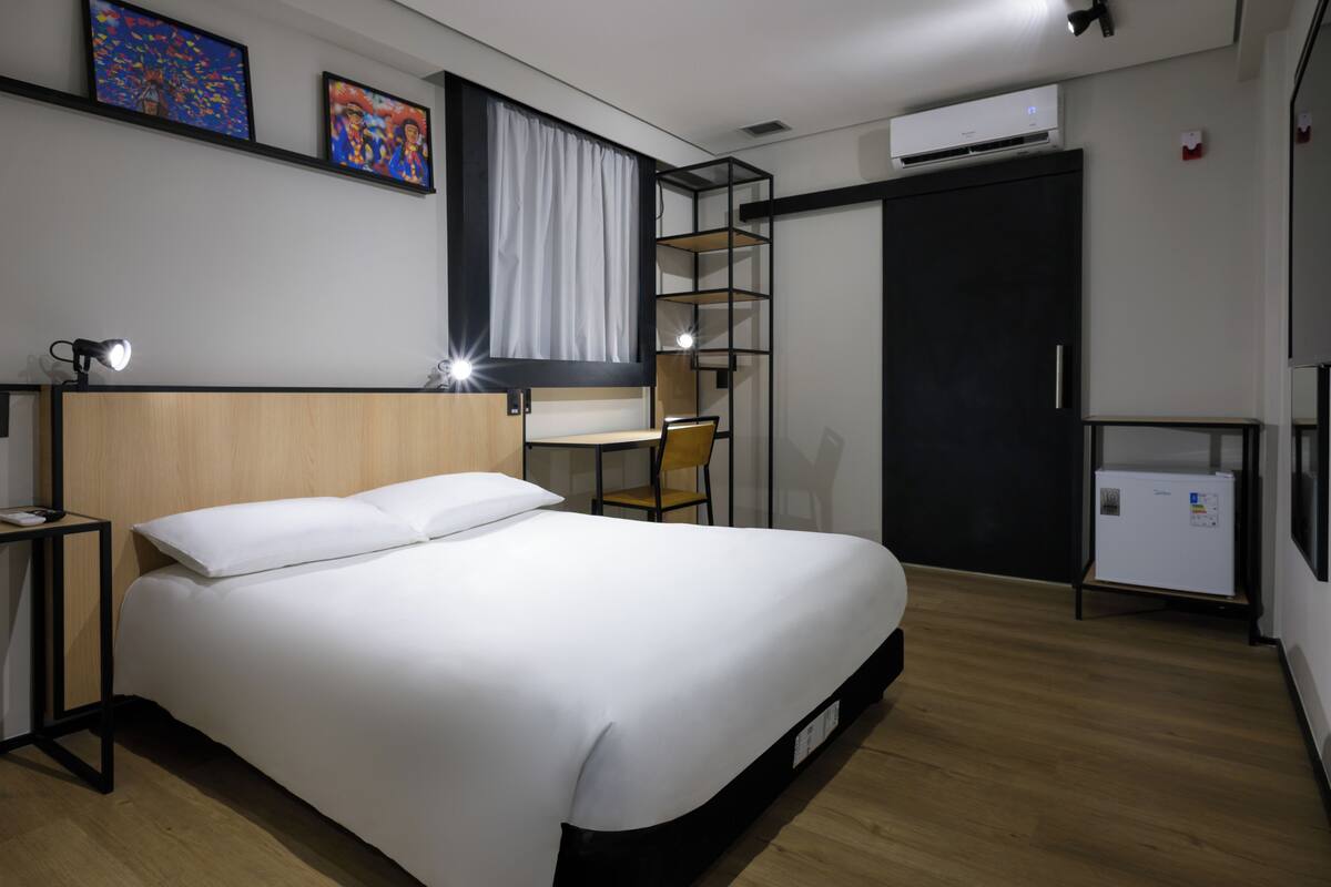 Ein modernes Hotelzimmer mit einem großen Bett, einem Schreibtisch, einem Sessel, einem kleinen Kühlschrank und Bildern an der Wand.