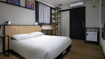 A modern hotel room with a large bed, a desk, a chair, a small refrigerator, and artwork on the walls.