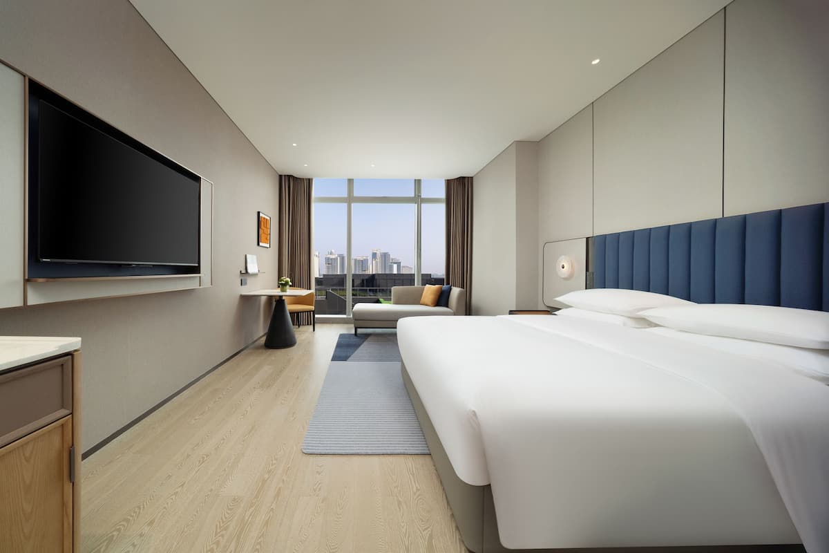 Ein modernes Hotelzimmer mit einem großen Bett, einem flachbildfernseher an der Wand, einer kleinen Sitzecke mit Sofa und Tisch sowie Blick auf die Stadtkulisse durch große Fenster.