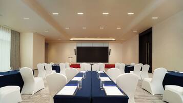Un salón de conferencias con una larga mesa, sillas blancas y una pantalla para proyector.