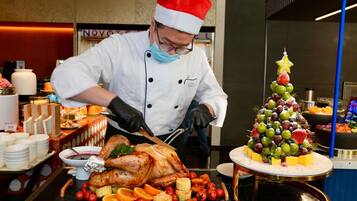 Un chef, coiffé d’un chapeau de fête, découpe une dinde rôtie devant une table de service où sont disposés divers fruits et légumes.
