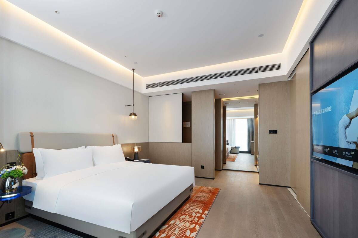 Una habitación de hotel moderna con una cama grande, mesitas de noche, un televisor y vistas al pasillo.
