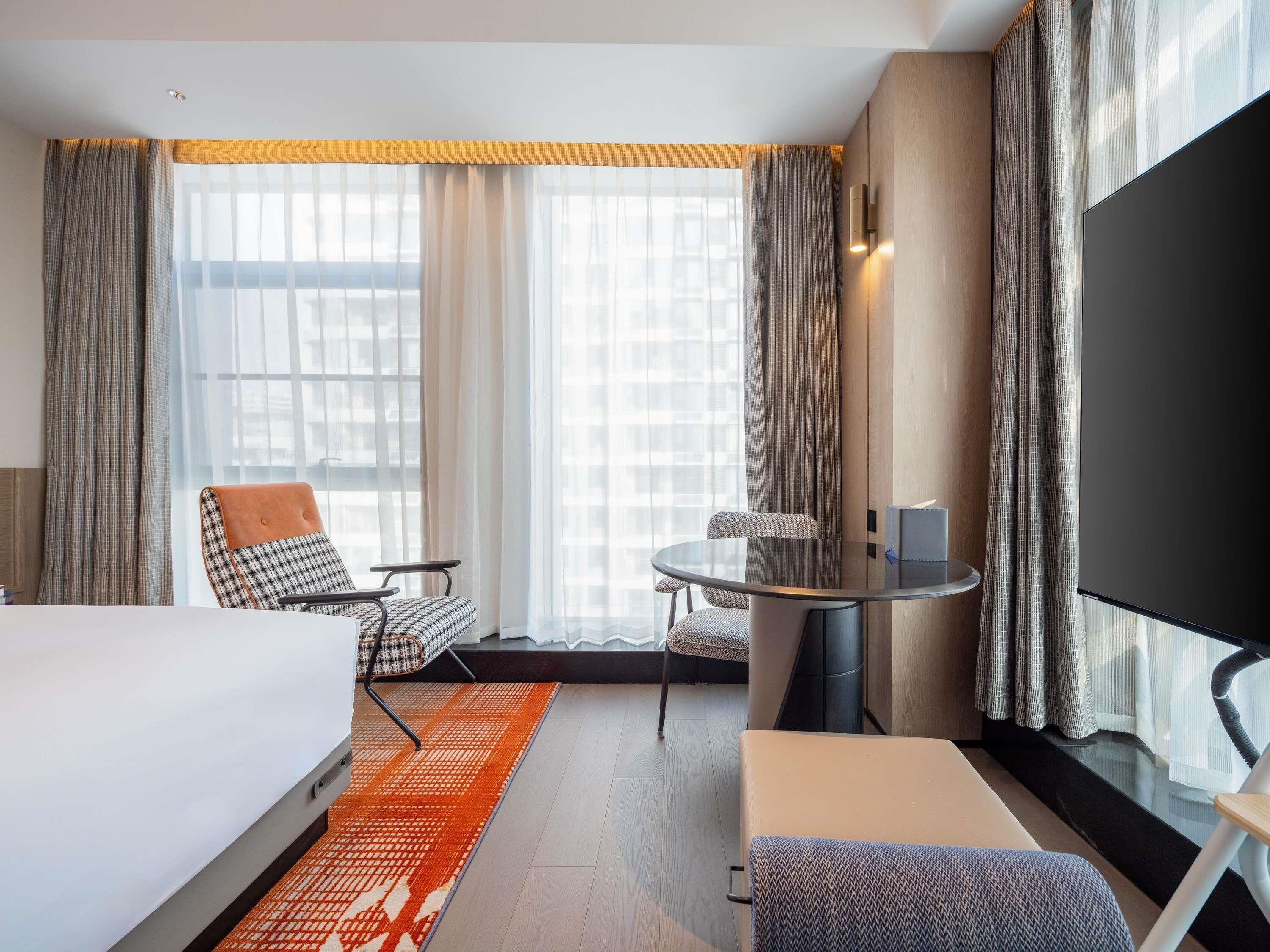 Quarto de hotel com cama, duas cadeiras, mesa pequena, televisão e janelas amplas com cortinas.