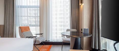 Quarto de hotel com cama, duas cadeiras, mesa pequena, televisão e janelas amplas com cortinas.