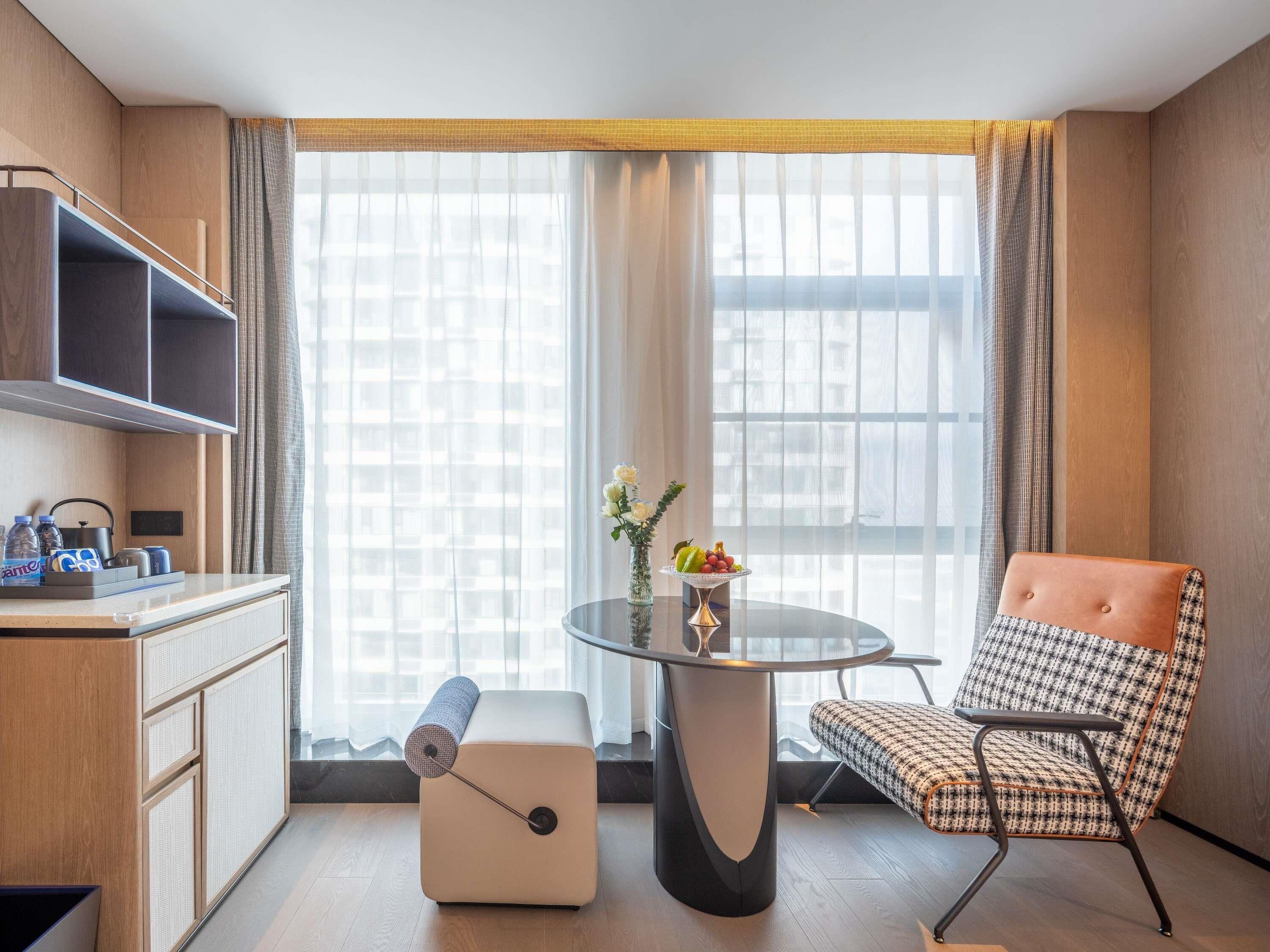 A compact hotel room with a kitchenette, a round table with a vase of flowers, a patterned armchair, and large windows with sheer curtains.
