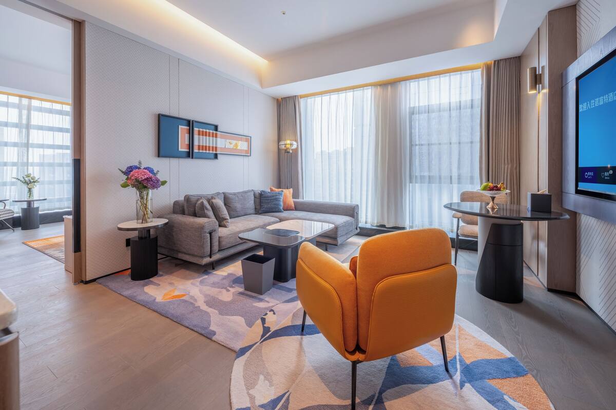 Una sala de estar moderna con sofá, mesa de centro y sillón naranja. Hay un televisor de pantalla plana montado en la pared, una mesita con fruta y un jarrón con flores.