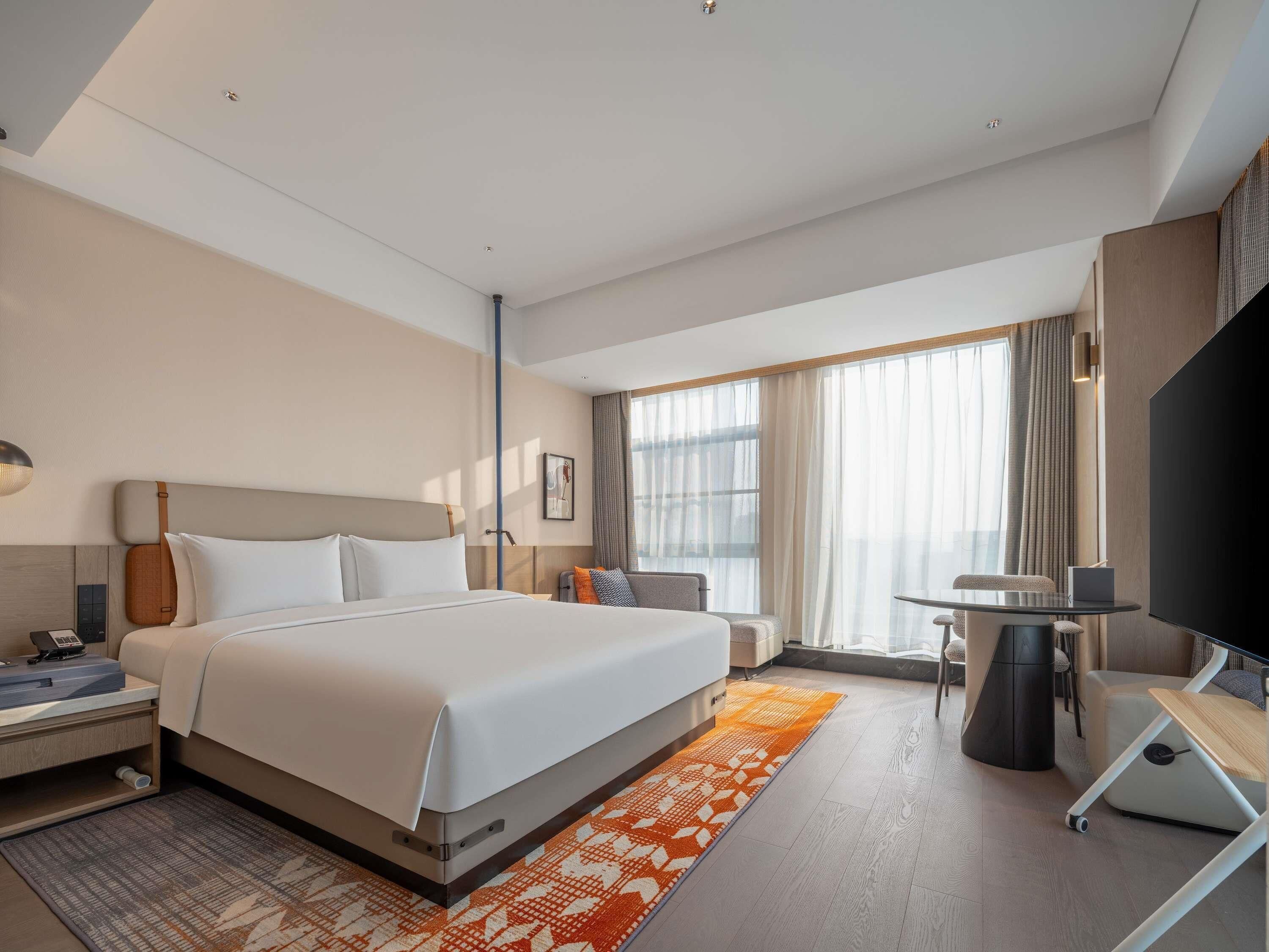Una habitación de hotel moderna con una cama grande, un escritorio con silla, un televisor de pantalla plana y un ventanal con cortinas.