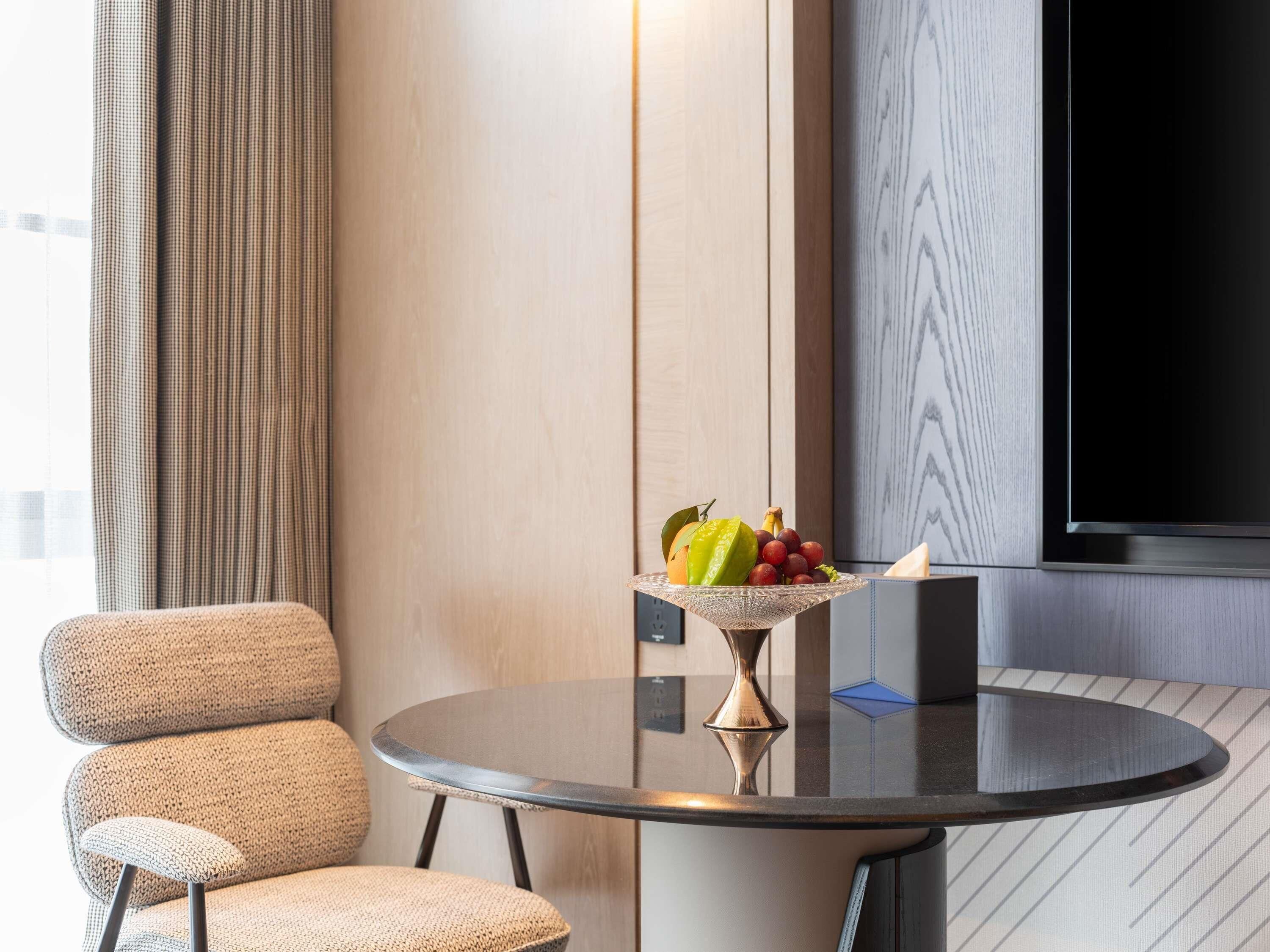 A modern hotel room with a round table, a chair, a fruit bowl, and a flat-screen TV.