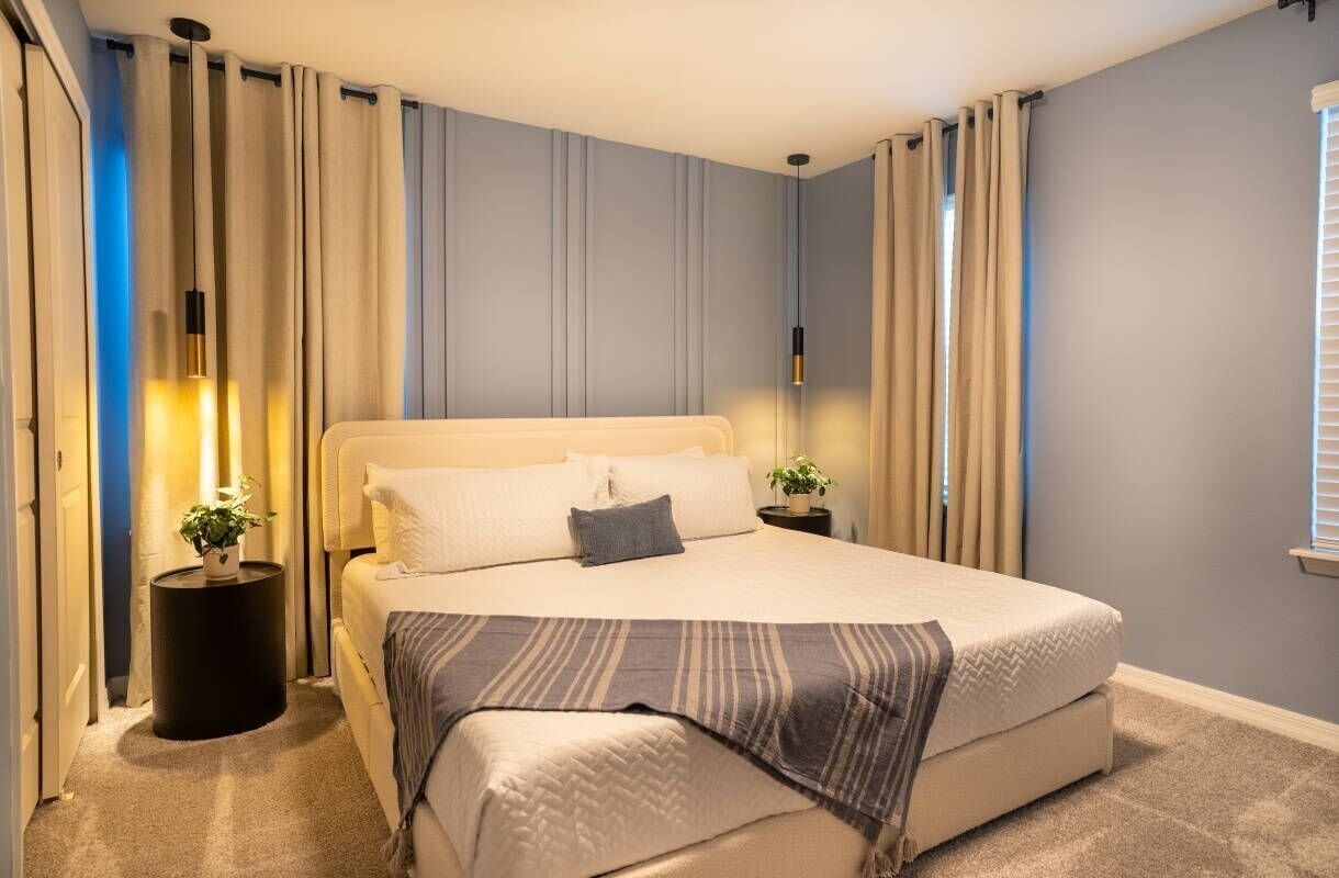Une chambre à coucher moderne avec un grand lit, deux tables de chevet, une plante en pot et une fenêtre avec des rideaux.