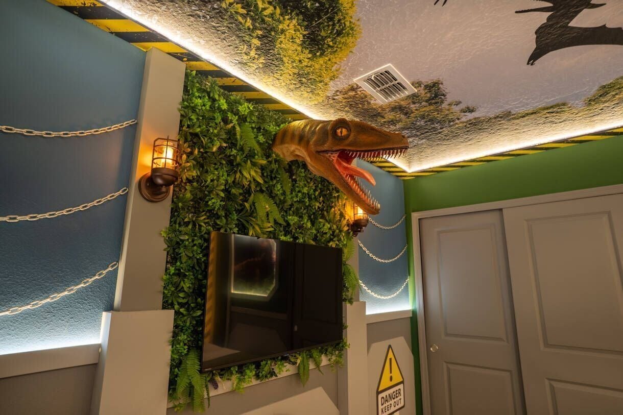 Una stanza con TV a parete, parete ricoperta di verde e decorata con una testa di dinosauro e un cartello di avvertimento.