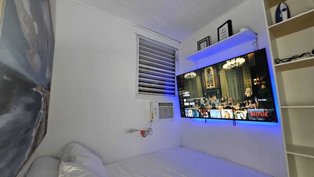 Une chambre à coucher avec un lit, un téléviseur fixé au mur, une fenêtre avec des stores et une étagère sur laquelle sont posés divers objets.