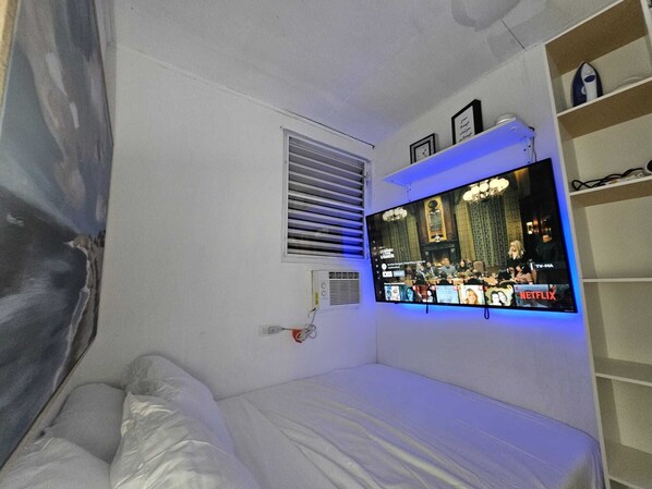 A bedroom with a bed, a TV mounted on the wall, a window with blinds, and a shelf with items.