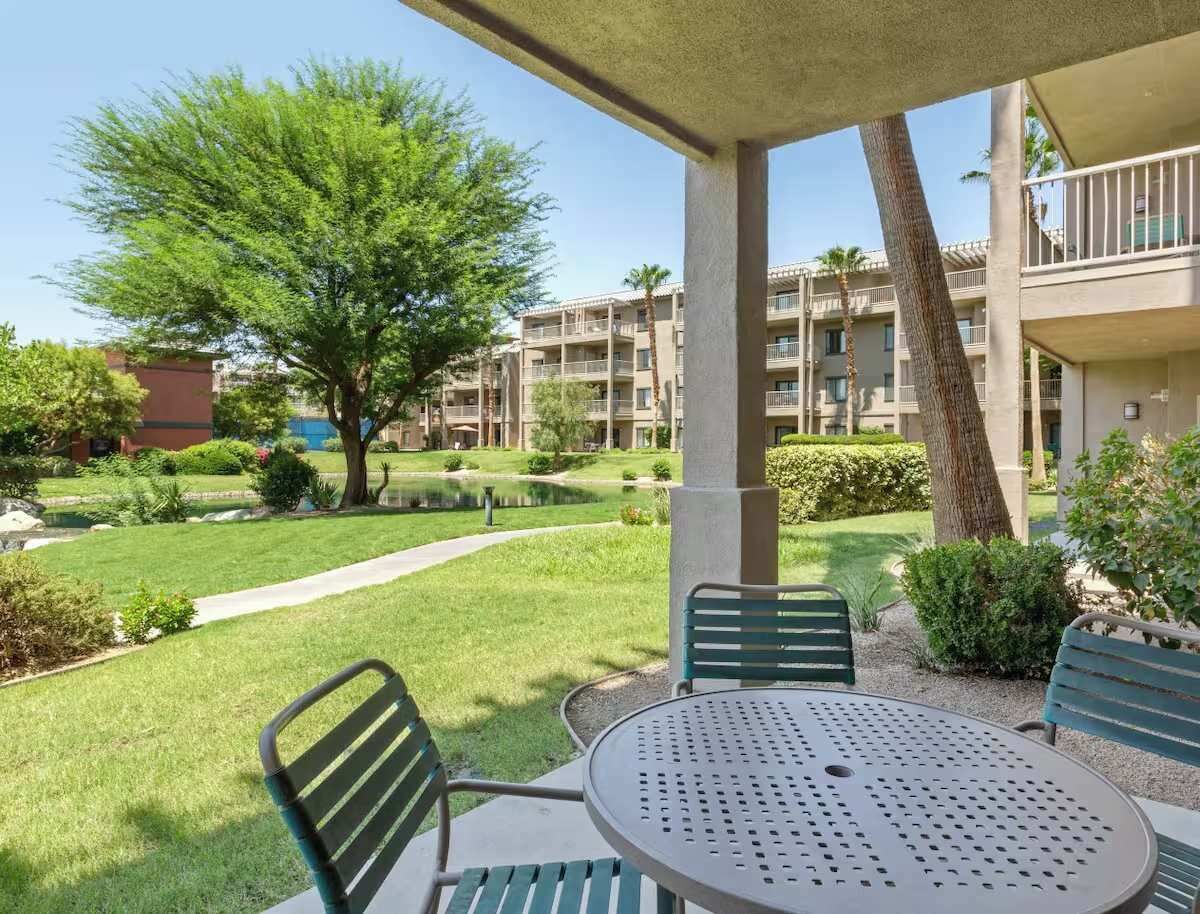 A courtyard with a round table and chairs, surrounded by greenery and a building with balconies.