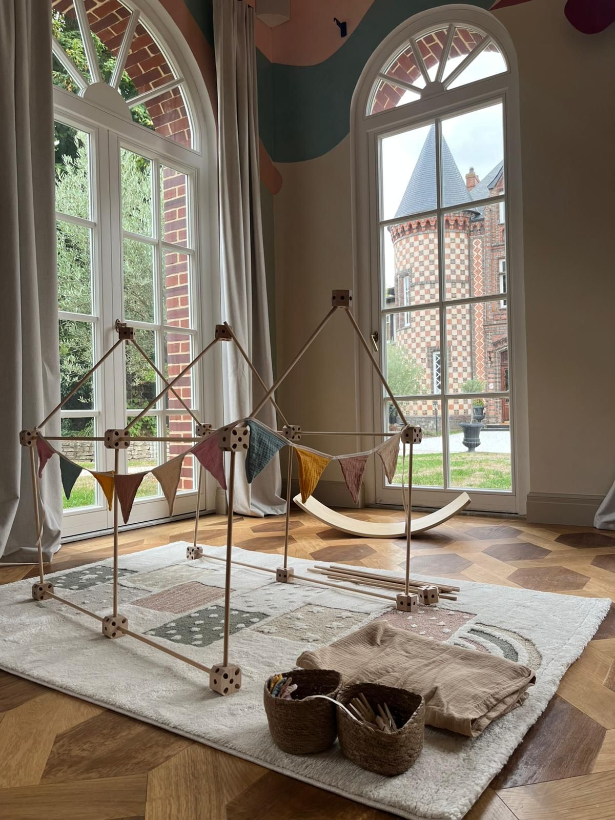 A room with large windows, a hammock, a wooden structure with triangular flags, and a patterned rug.