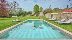 Piscina rectangular con diseño ajedrezado, rodeada por un muro de piedra, con tumbonas y sombrillas.