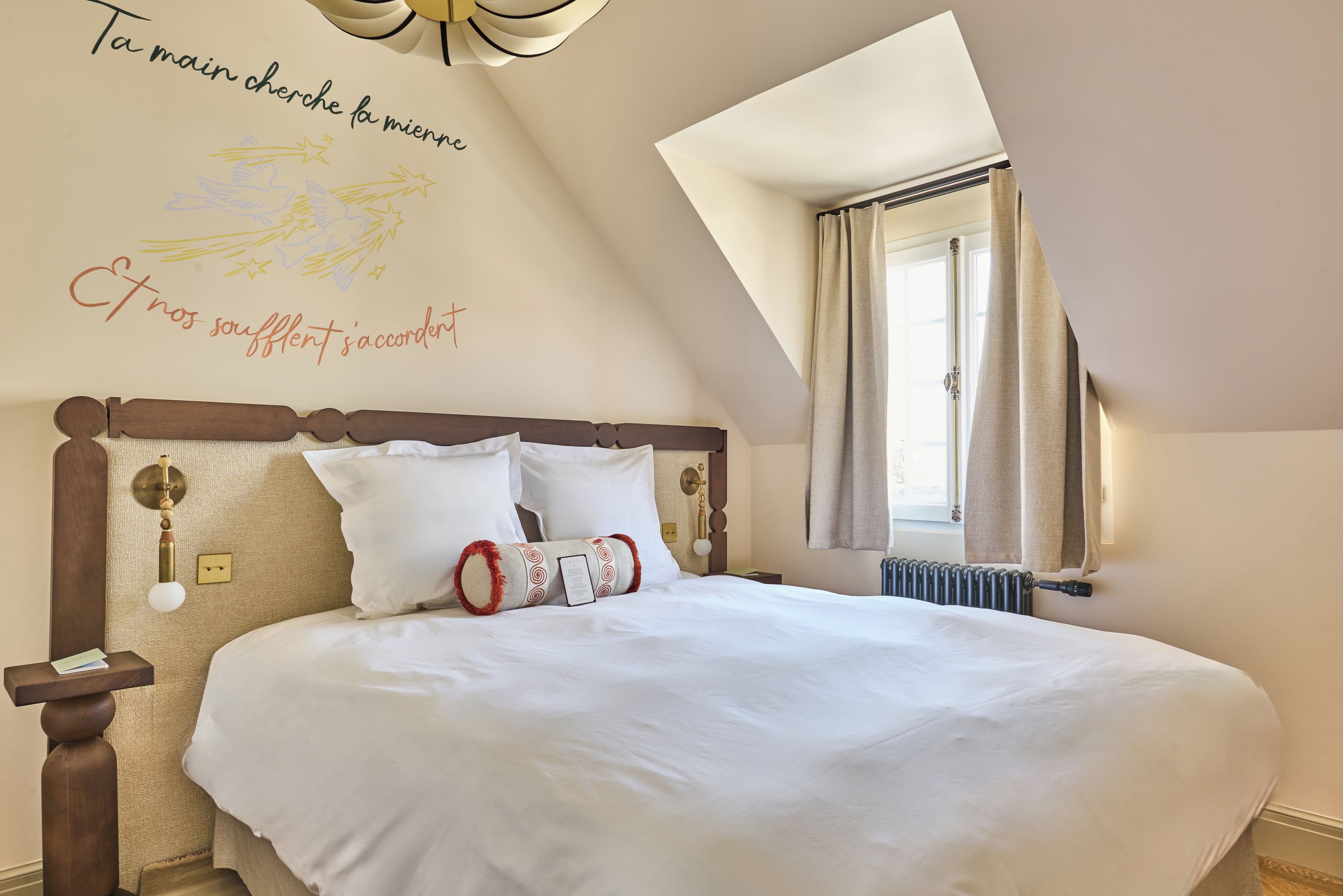 A bedroom with a large bed, a headboard with wall-mounted lights, a window with curtains, and a wall with decorative text and star drawings.