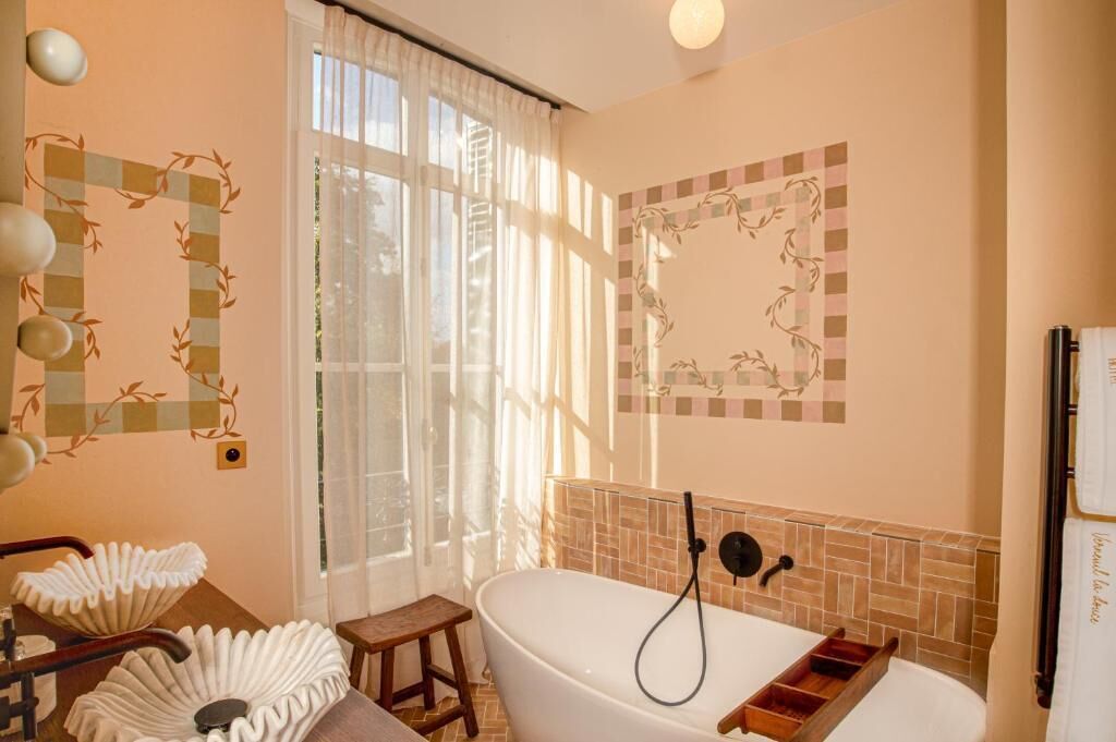 Ein Badezimmer mit freistehender Badewanne, einem Waschtisch mit Spiegel und einem Fenster mit transparenten Vorhängen.