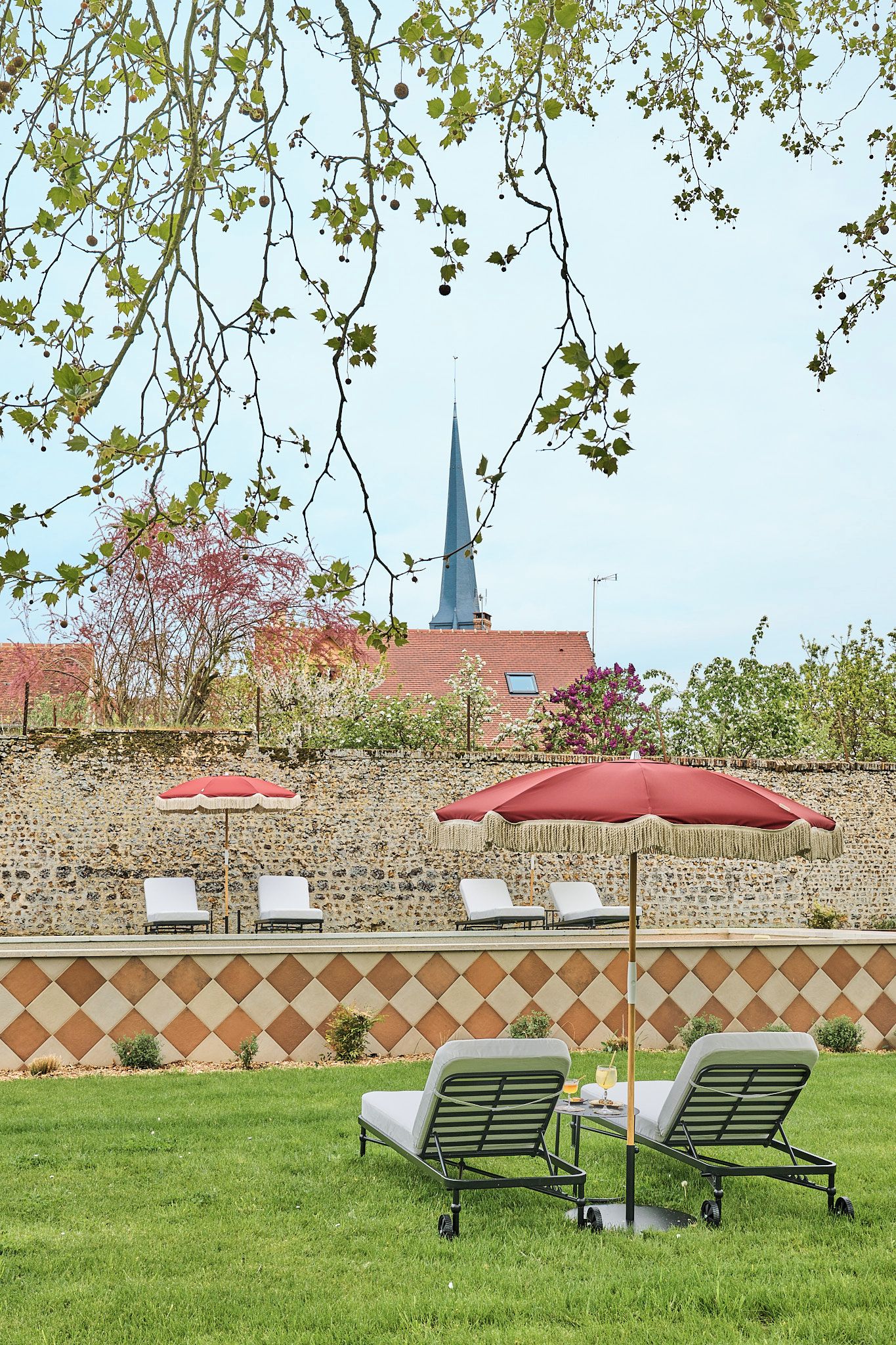 A patio area with lounge chairs and umbrellas, a stone wall, and a building with a spire in the background.