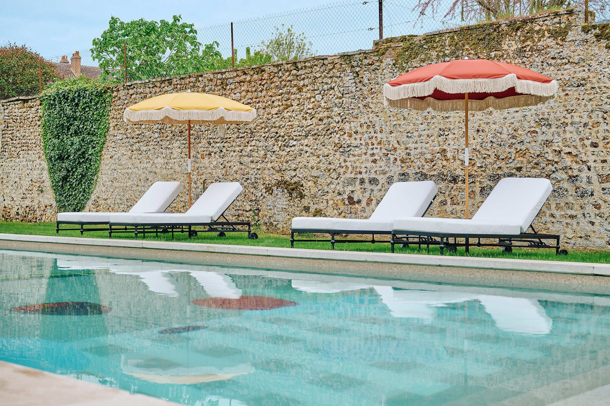 A swimming pool with lounge chairs and umbrellas, surrounded by a stone wall and greenery.