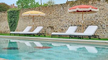 A swimming pool with lounge chairs and umbrellas, surrounded by a stone wall and greenery.