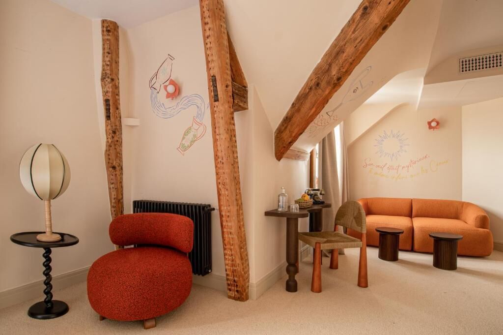 A room with exposed wooden beams, a red chair, an orange sofa, a small table, and a wall with a decorative mural.