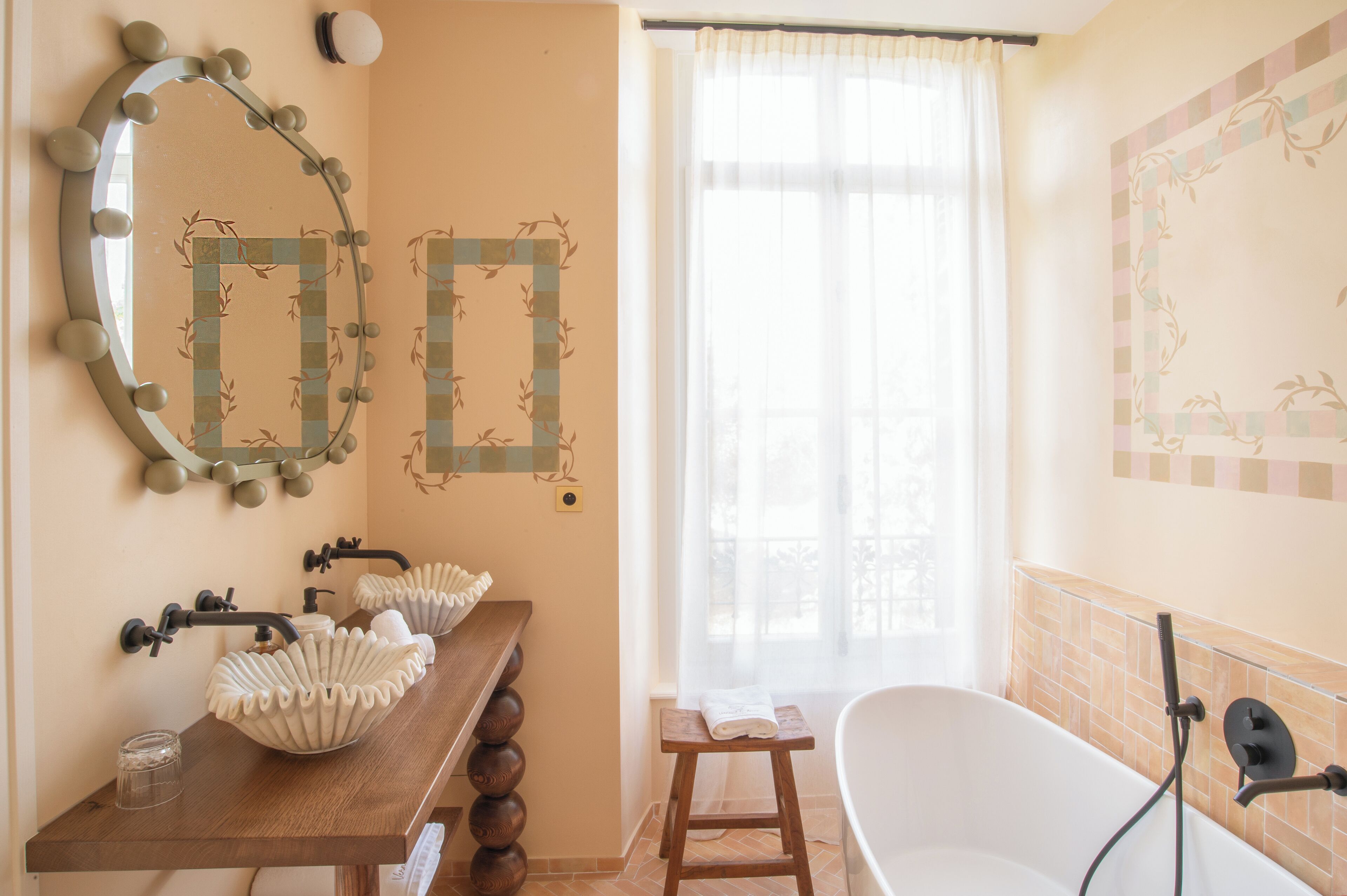 A bathroom with a vanity, two mirrors, a bathtub, and a window with sheer curtains.