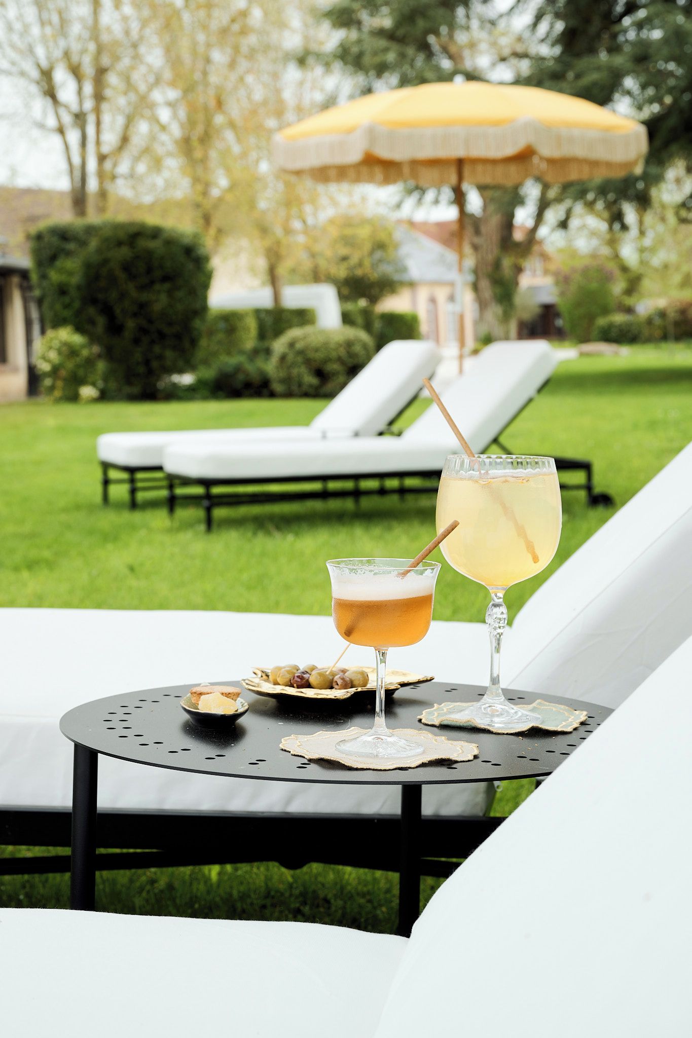 Ein Tisch mit zwei Getränken und einer Olivenplatte, vor einem Garten mit Sonnenschirm und Liegestühlen.