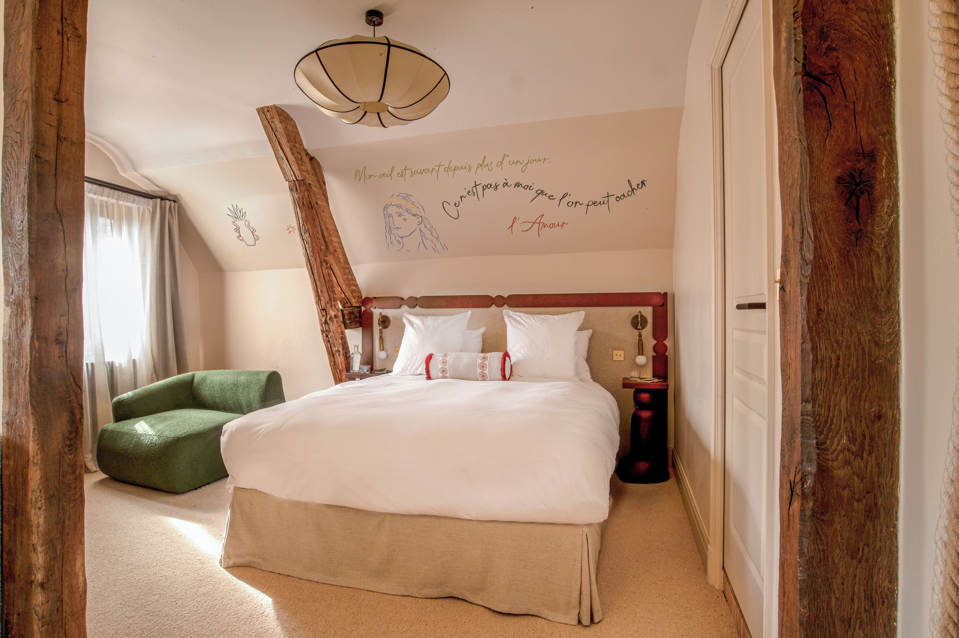 Una habitación de hotel con una cama grande, una silla verde, un cabecero de madera, una pared con una cita decorativa y una lámpara de techo.