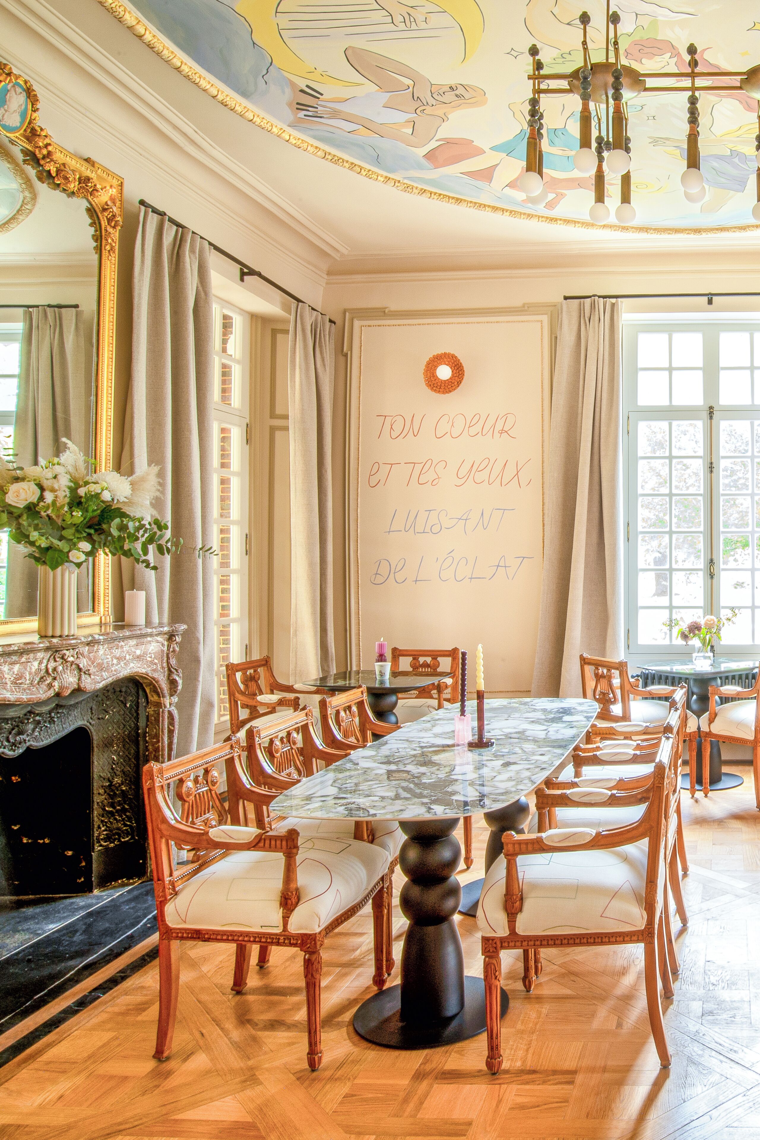 A dining room with a marble table, wooden chairs, a fireplace, and a mural on the ceiling.