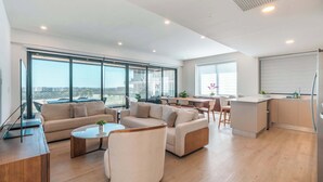 Een ruime woonkamer met een bank, fauteuils en een salontafel. Er is een keuken met een aanrecht en kasten. Grote ramen zorgen voor daglicht en uitzicht op het omliggende landschap.