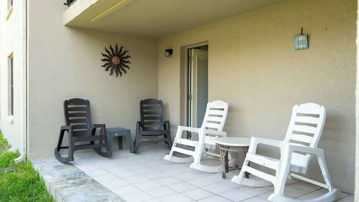 Un espace extérieur abrité avec quatre chaises et une petite table, une décoration murale en forme de soleil, et une porte donnant sur l’intérieur.