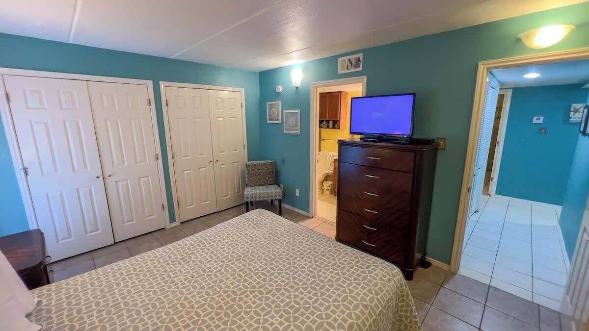 Une chambre à coucher comprenant un lit, une commode, un téléviseur et une porte donnant sur une autre pièce.