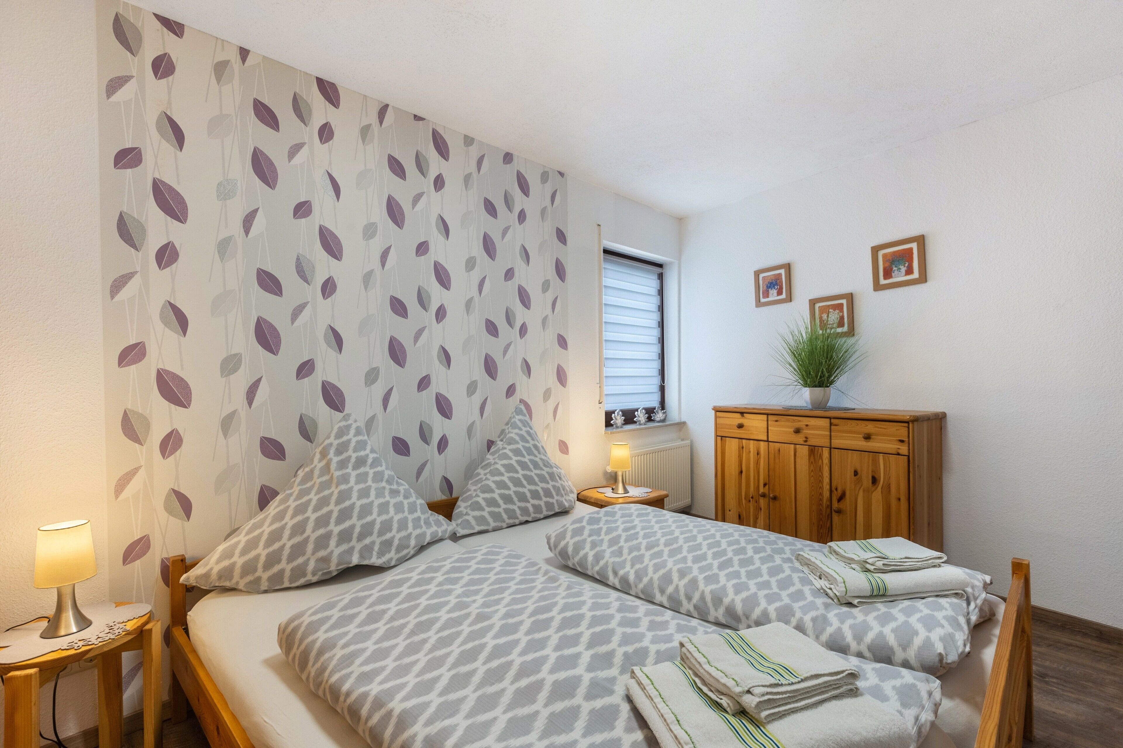 A bedroom with two beds, a nightstand with a lamp, a wooden cabinet, and a window with blinds.
