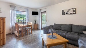 Een woonkamer met een bank, een houten tafel, een aan de muur bevestigde televisie, en een eethoek met stoelen en een tafel.