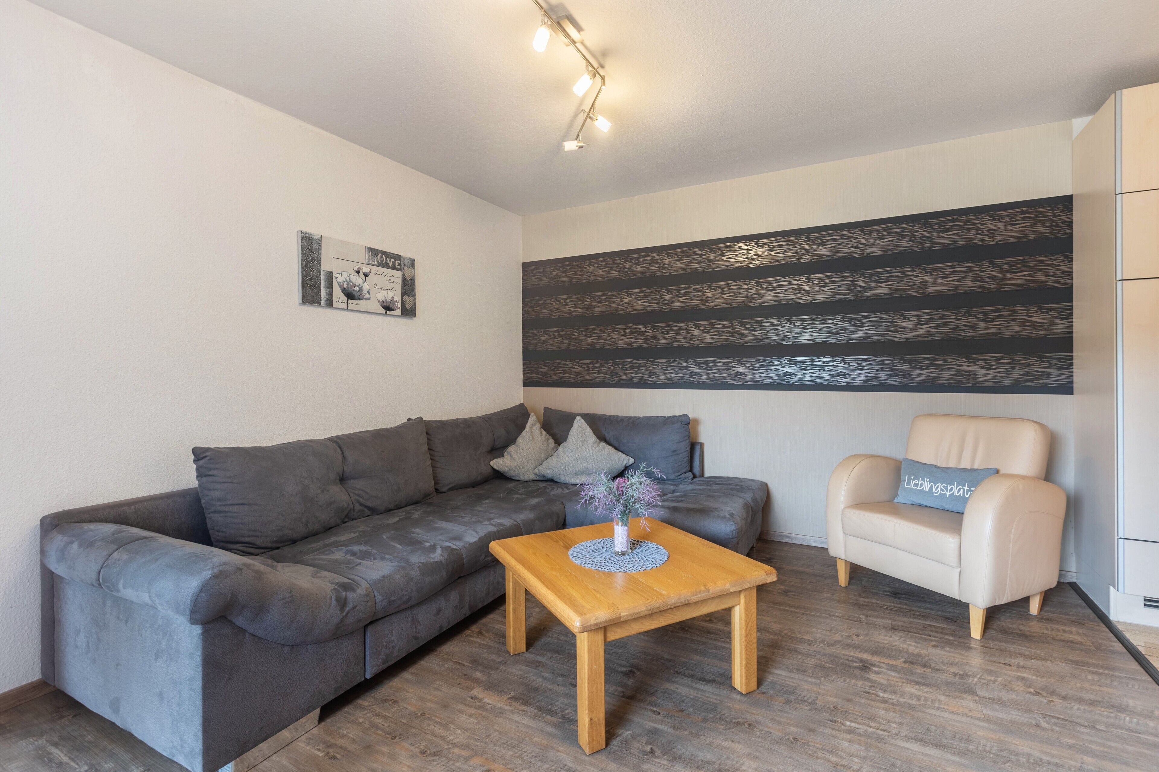 Un salon moderne avec un canapé gris, une table basse en bois, un fauteuil beige et une grande œuvre d’art abstraite accrochée au mur.