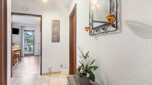 Een gang met een potplant, een spiegel met wandlampen en een houten deur die leidt naar een kamer met een tafel en stoelen.