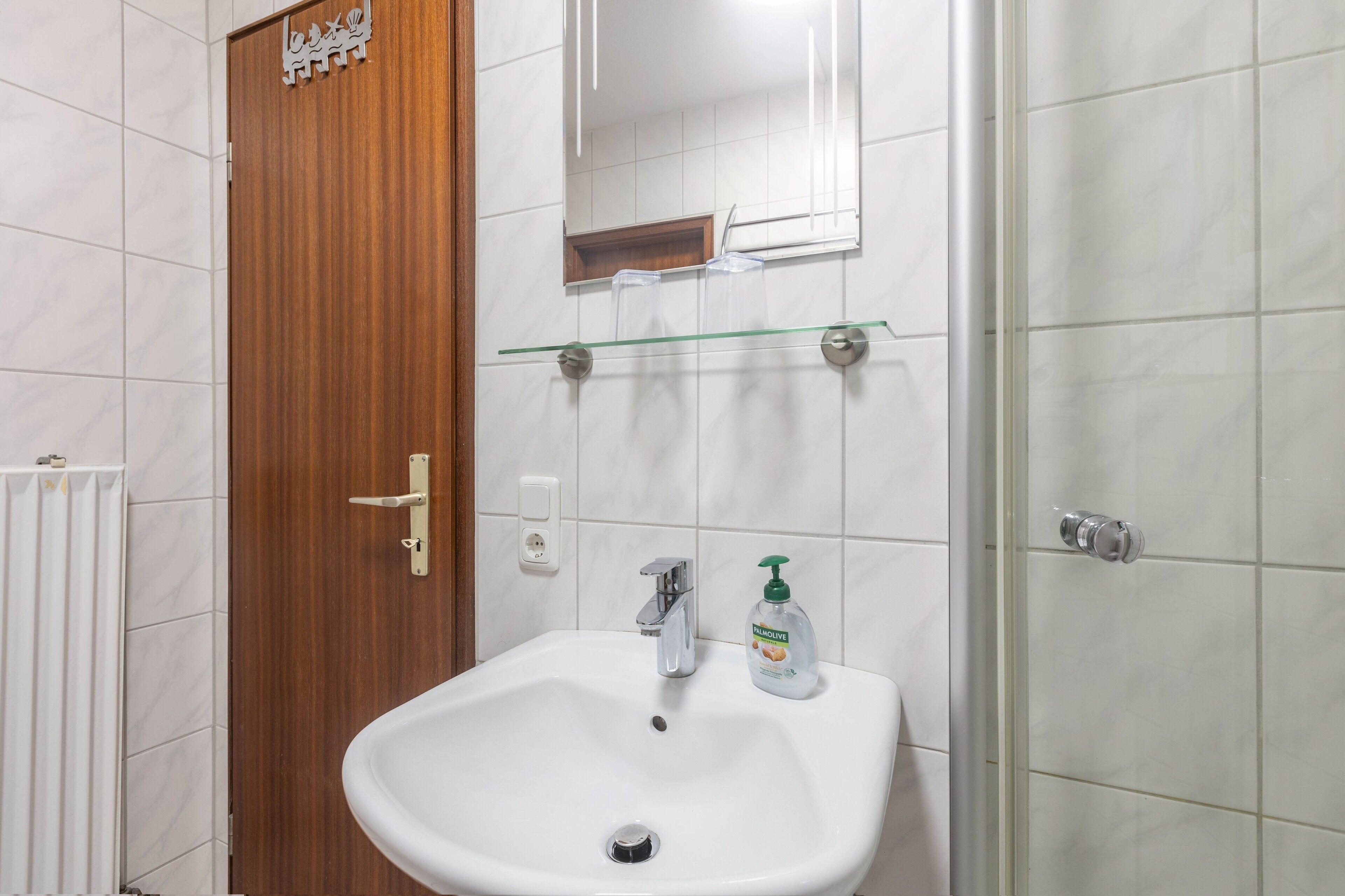 Een badkamer met een houten deur, een wit wastafelmeubel met een spiegel erboven en een douchehoek.