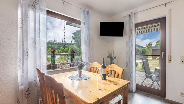 Een eetruimte met een houten tafel, stoelen, een aan de muur bevestigde televisie en uitzicht op een tuin door de ramen.