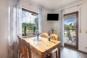 A dining area with a wooden table, chairs, a TV mounted on the wall, and a view of a garden through the windows. - Sauerland Apartment – Oasis with Private Terrace, Garden, and Wi-Fi (Medebach)