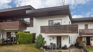 Een modern huis met twee verdiepingen, een balkon, een klein terras met tuinmeubilair en een verzorgde tuin.