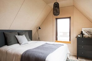 Et soveværelse med skrå væg, en seng med gråt tæppe, en mørk kommode og et vindue med naturligt lys.