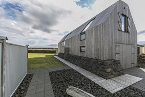 Un edificio moderno de madera con techo inclinado, base de piedra y un sendero pavimentado que conduce a una zona verde.