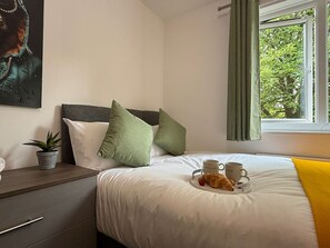 Ein ordentlich bezogenes Bett mit zwei weißen Kissen, einem grünen Kissen und einer gelben Decke. Ein Tablett mit Croissants und zwei Tassen steht auf dem Bett.