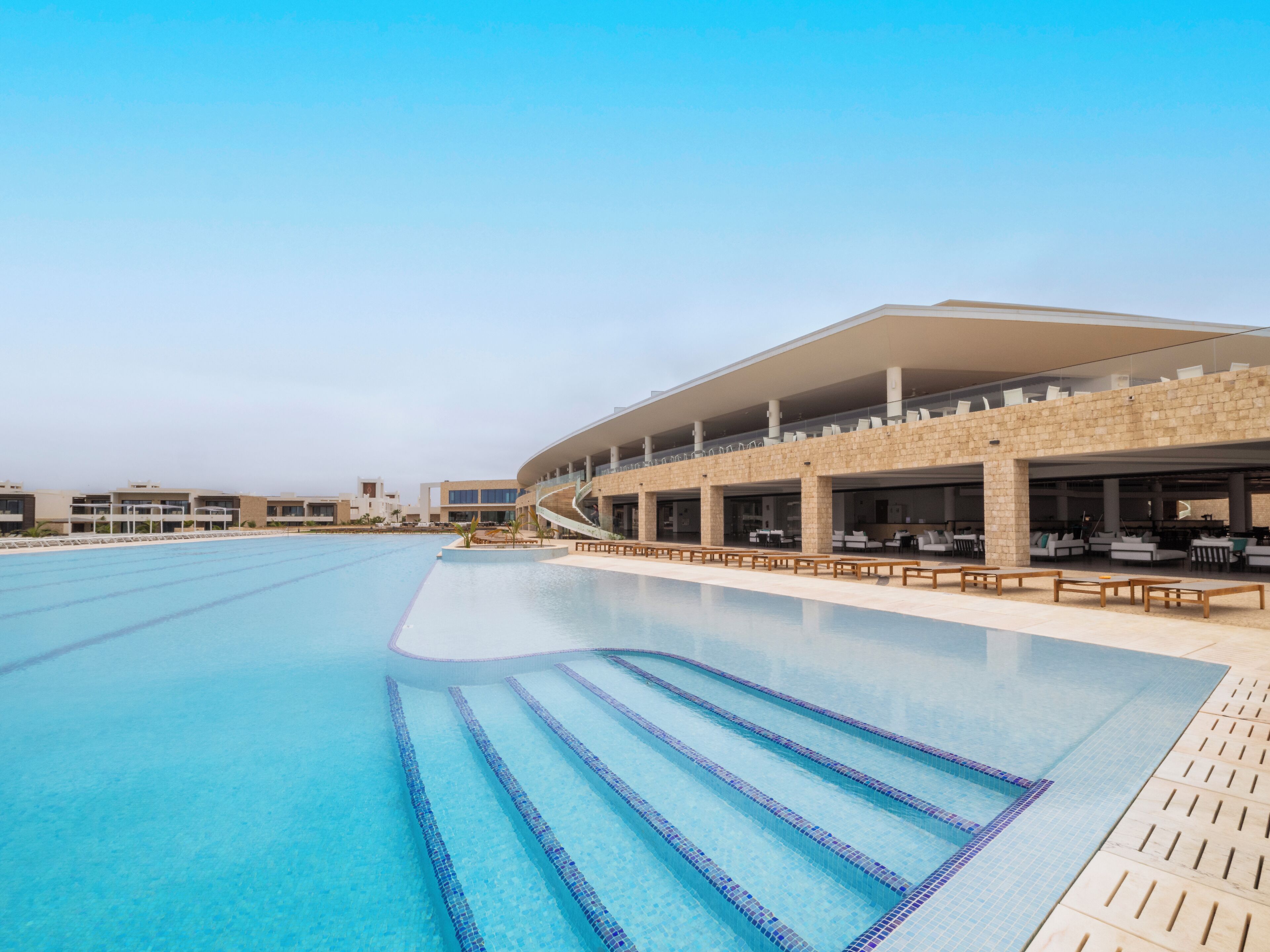 Una piscina con agua azul cristalina, una zona de estar techada con mobiliario exterior y un edificio moderno con techo curvo.