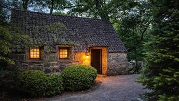 Una cabaña de piedra con techo de paja, puerta de madera y dos ventanas con luz cálida.