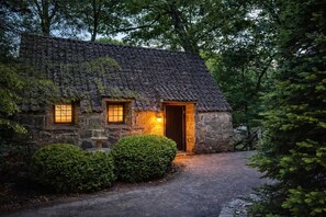 A stone cottage with a thatched roof, a wooden door, and two windows with warm lighting. - Storybook 3-Cottage Mountain Getaway (Banner Elk)